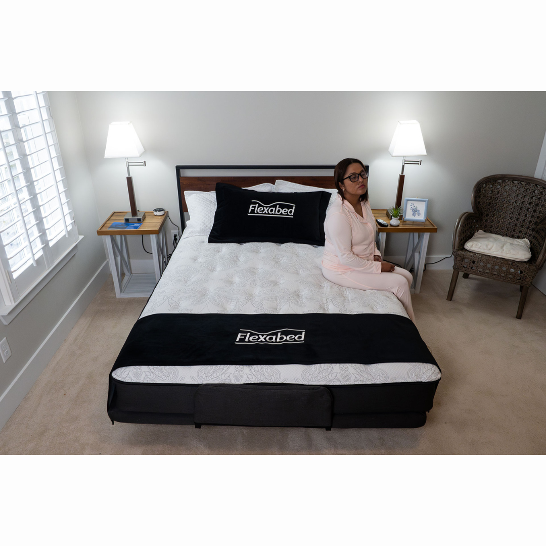 A person in pajamas sits on the edge of a Flexabed Premier Adjustable Full Electric Bed with Massage in a softly lit bedroom. The room includes customizable configurations like a nightstand with a lamp and a decorative chair, while the bedding displays Flexabed branding.